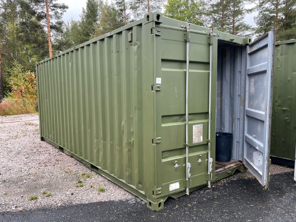 A green container with an open door on a sandy field. Asphalt in front and forest behind.