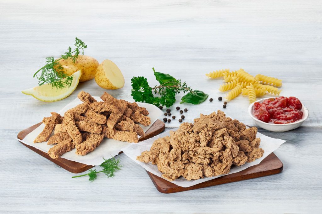 Strips and shreds of small fish on wooden boards. Lemon slices, potatoes, herbs, pasta, and ketchup in a bowl in the background. 