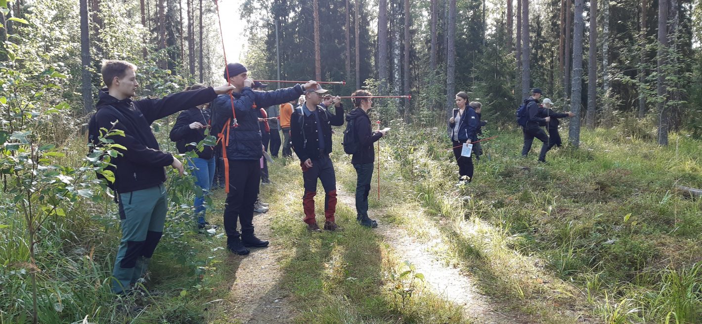 Students stand in the forest with measuring sticks.