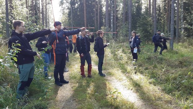 Students stand in the forest with measuring sticks.